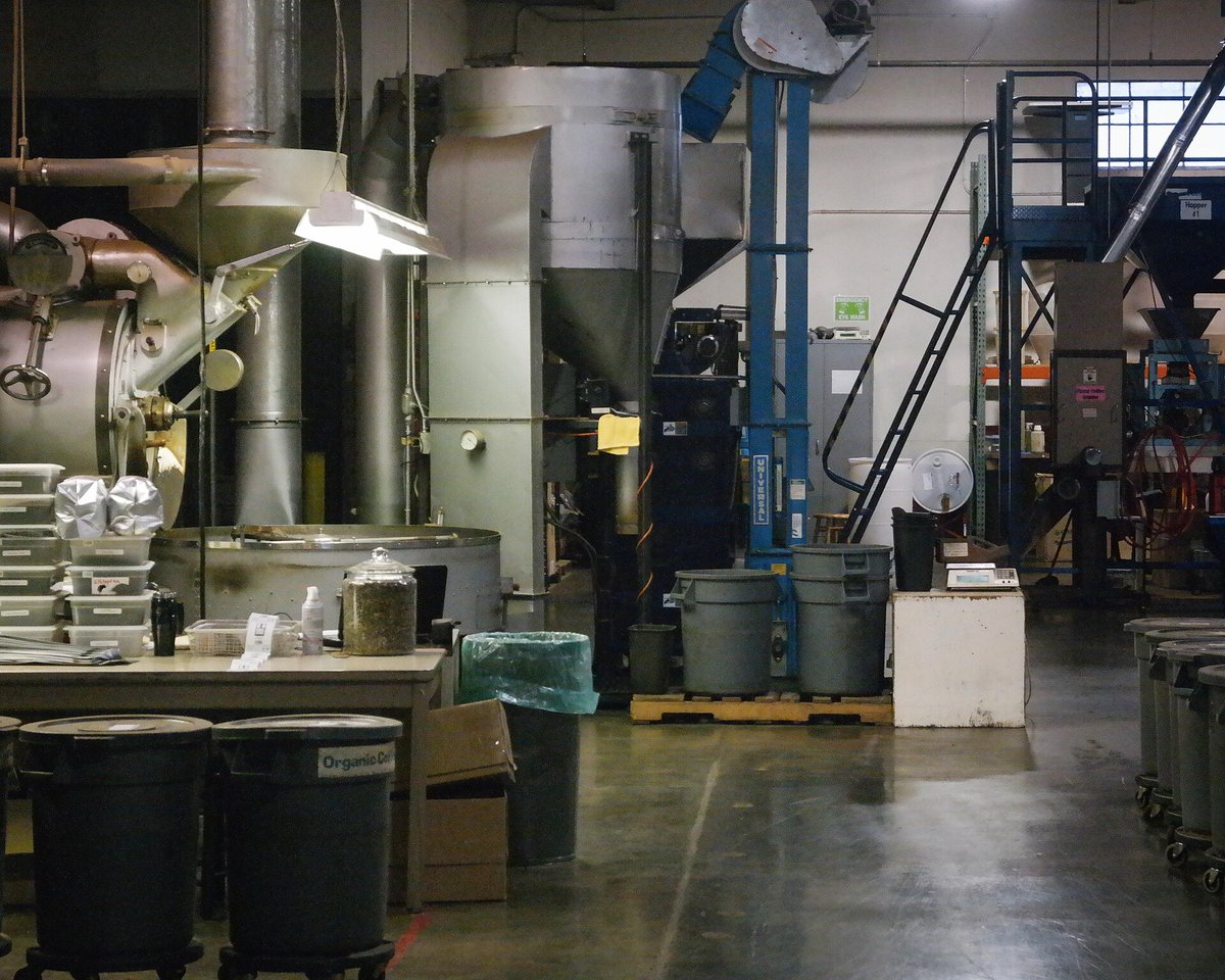 A production roasting room with a large drum roaster, ducting to the ceiling, bins and sacks on a concrete floor