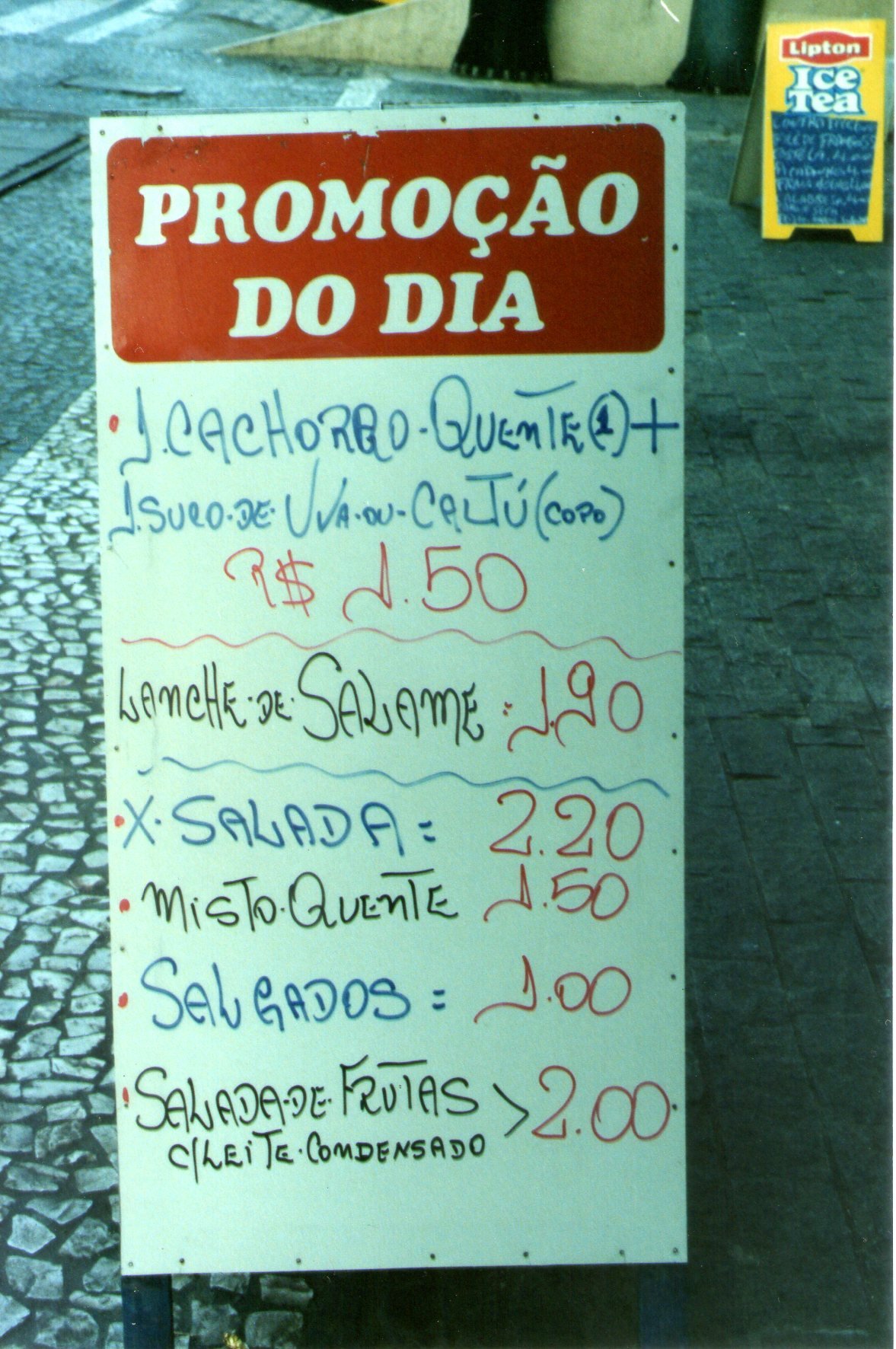 A handwritten street menu board in a plastic sleeve, headed Promoção do dia, listing snacks and prices in blue and red marker