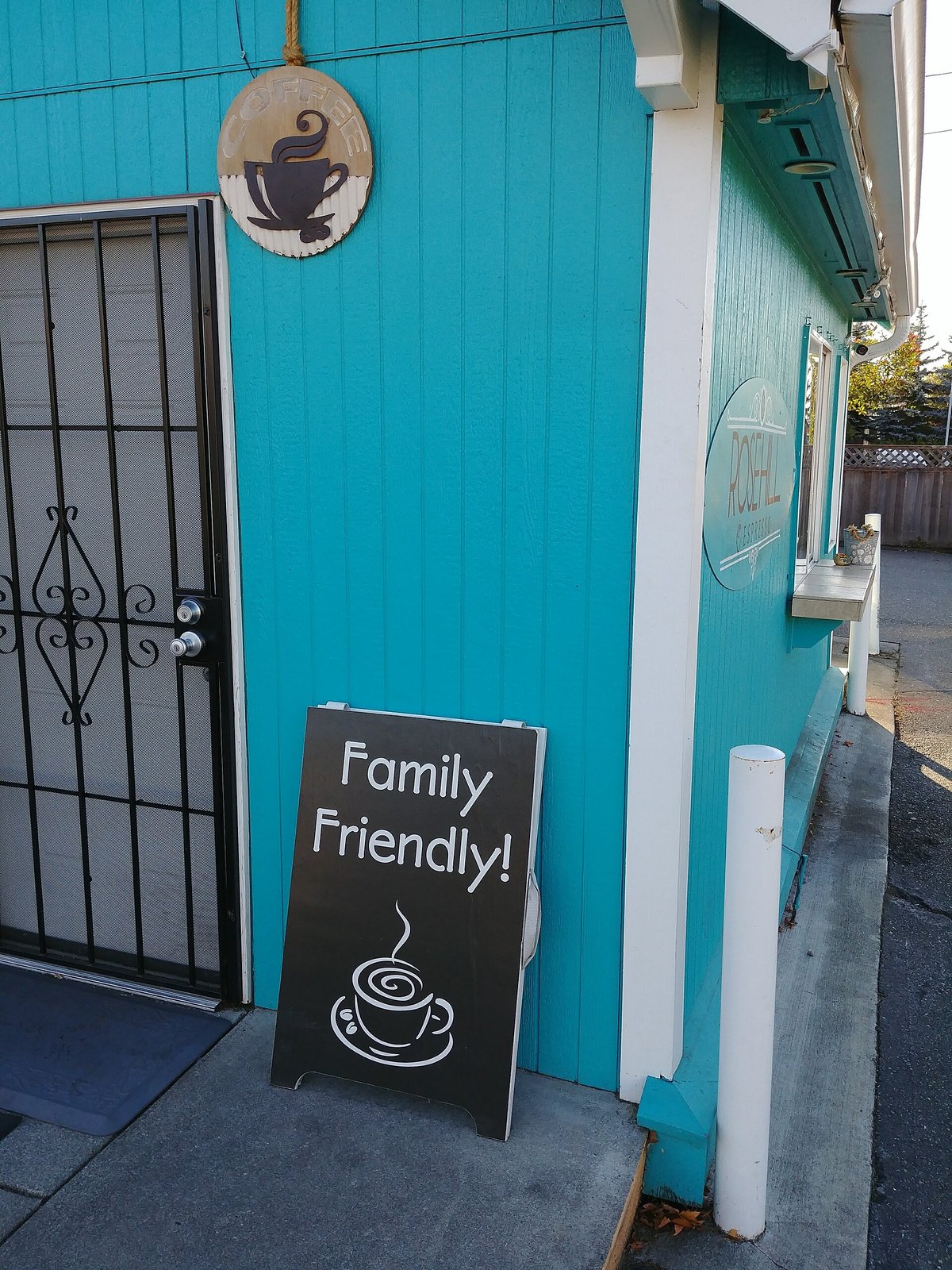 A small teal drive-through coffee stand with a sandwich board beside it reading Family Friendly with a drawing of a steaming cup