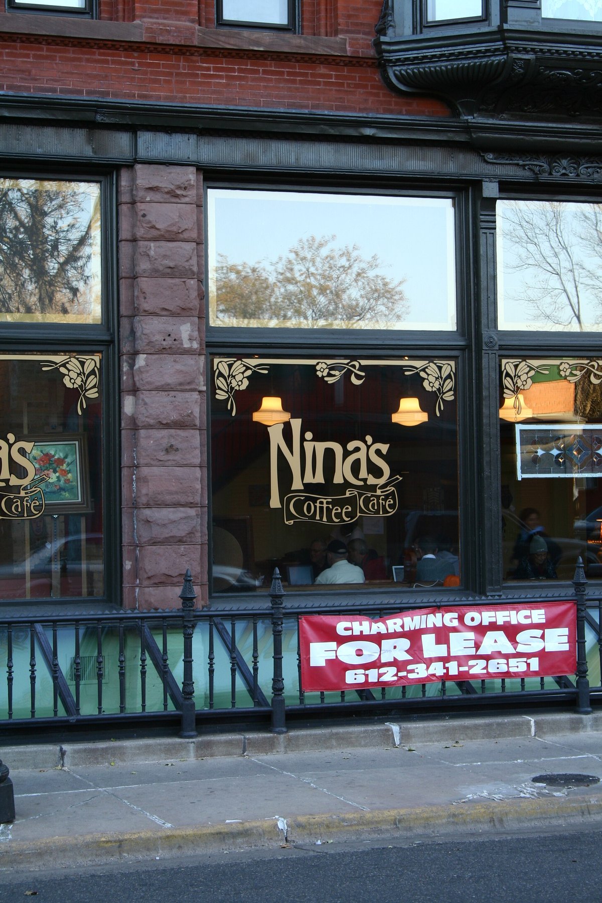 A brick storefront with arched windows lettered Nina's Coffee Café, and a red For Lease sign hung across the railing in front