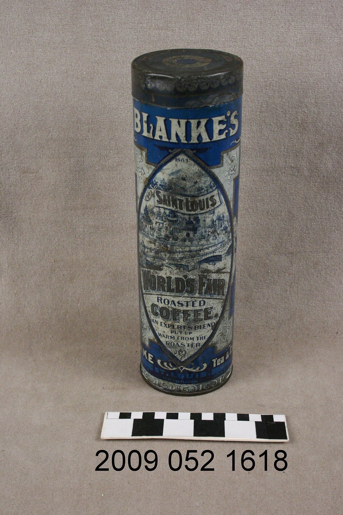 A tall cylindrical tin, blue with silver lettering reading Blanke's, with a museum catalogue number and a black-and-white scale bar in front of it