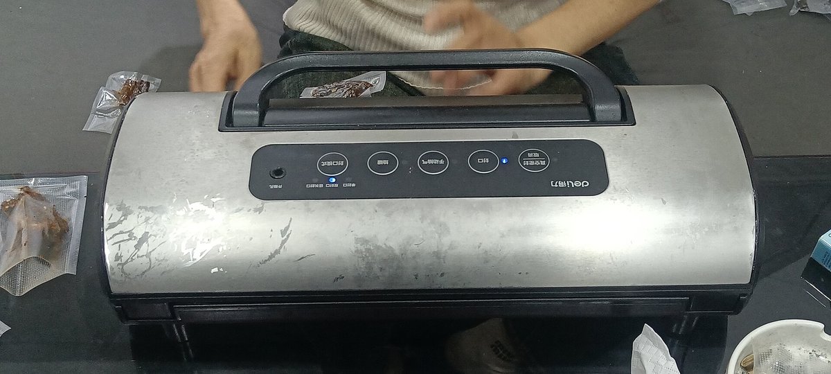 A silver bar-type vacuum sealer on a table with a hand resting on the lid and a plastic bag clamped under it