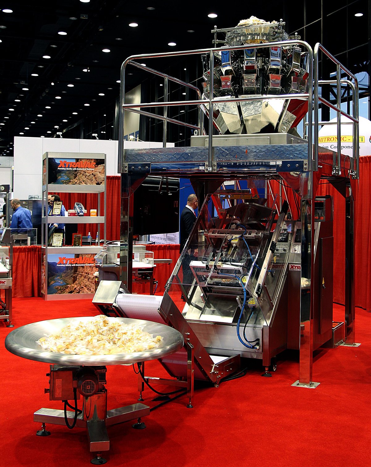 A tall stainless-steel packaging machine at a trade show: a multihead weigher on top drops product down into a vertical form-fill-seal machine, which forms bags from a roll of film and drops them onto a round catching table