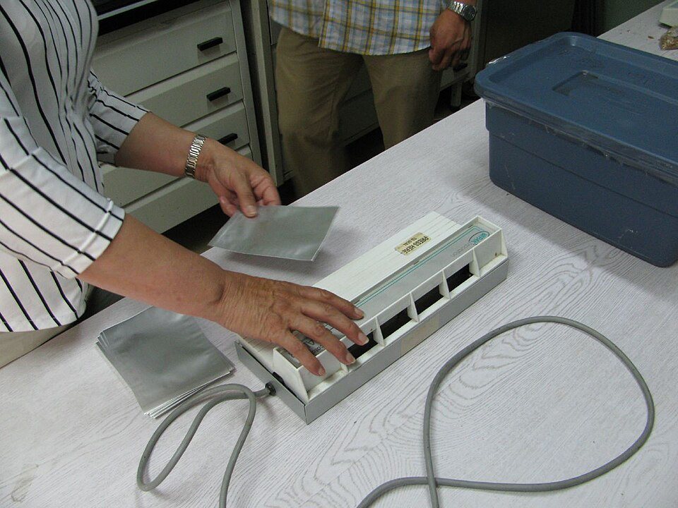 Two hands feeding a flat silver foil pouch into a tabletop heat sealer on a workbench, more foil pouches stacked beside it