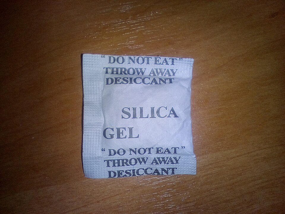 A small white paper sachet printed Silica Gel, do not eat, throw away, desiccant, lying on a wooden table