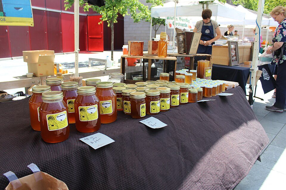 Jars of honey in rows on a dark tablecloth at an outdoor market, a vendor in an apron behind