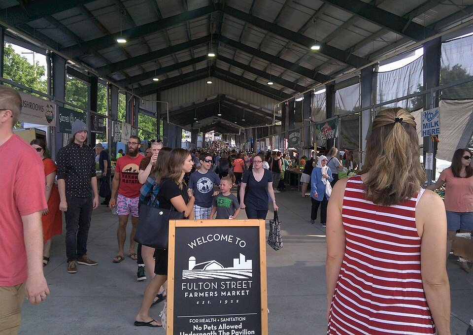 A dense crowd of shoppers under the roof of a covered market aisle, beside a sign reading Welcome to Fulton Street Farmers Market