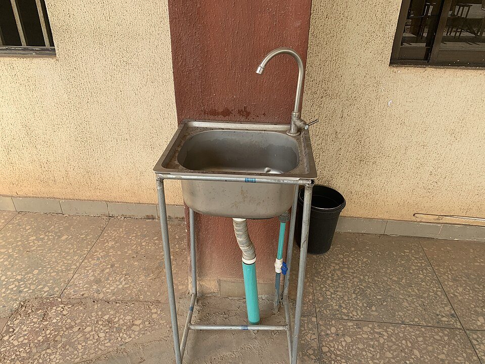 A small stainless steel sink on a metal frame standing against an outside wall, with a bucket beside it