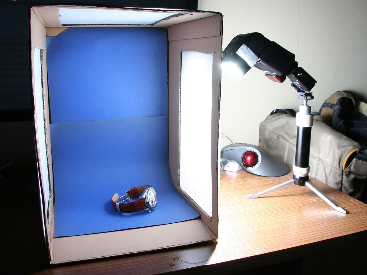 A homemade light box made of cardboard with white paper on the sides, a blue backdrop, a lamp on a small tripod and a small object being photographed
