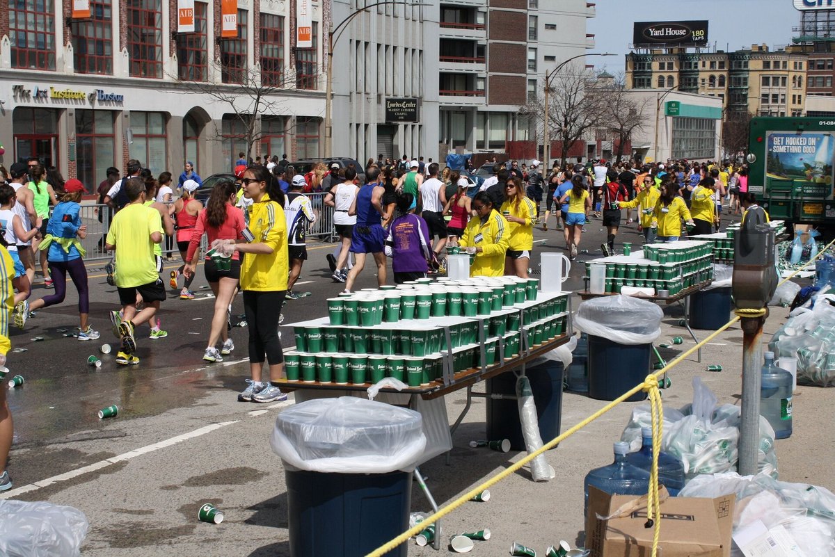 A street crowd of runners passing a long table stacked with cups of water during a marathon, volunteers behind it, discarded cups on the ground