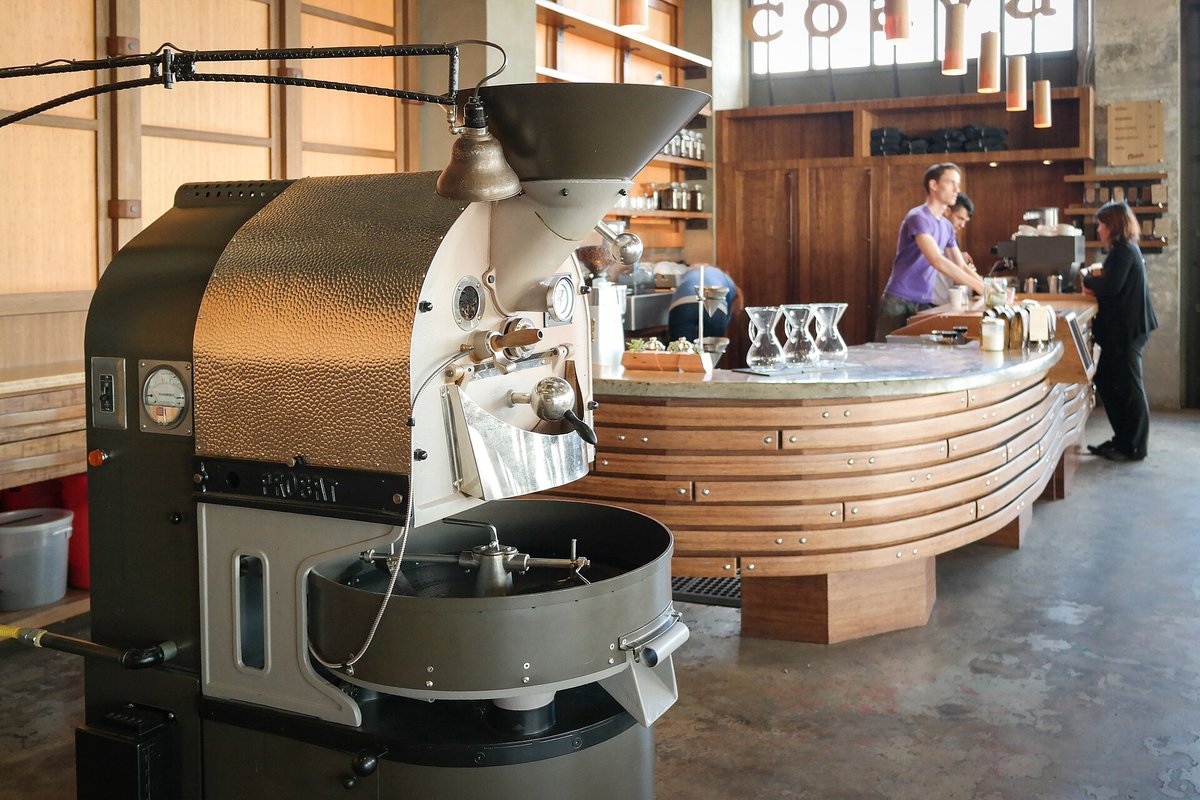 A shop drum roaster with a hopper and cooling tray in a café, a long curved wooden counter behind it and two people at the bar