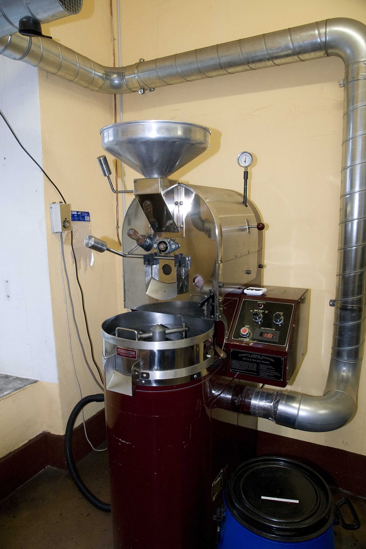 A small drum roaster in the corner of a room, with a wide spiral steel duct running from the back of the machine up the wall and across the ceiling, and a cooling tray in front