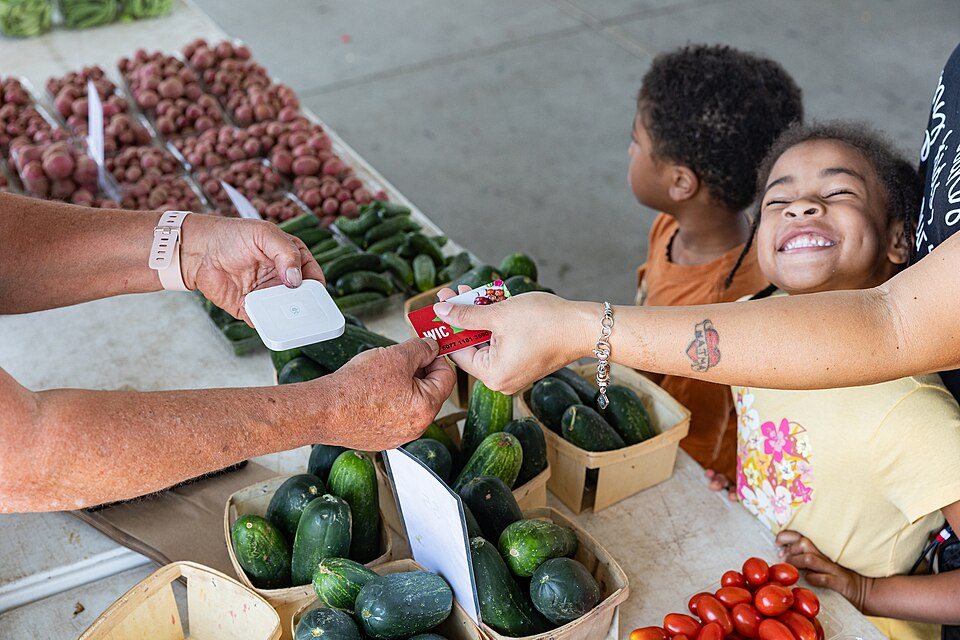 Over a table of cucumbers, a shopper hands a card to a vendor who holds a small white card reader, a smiling child looking on