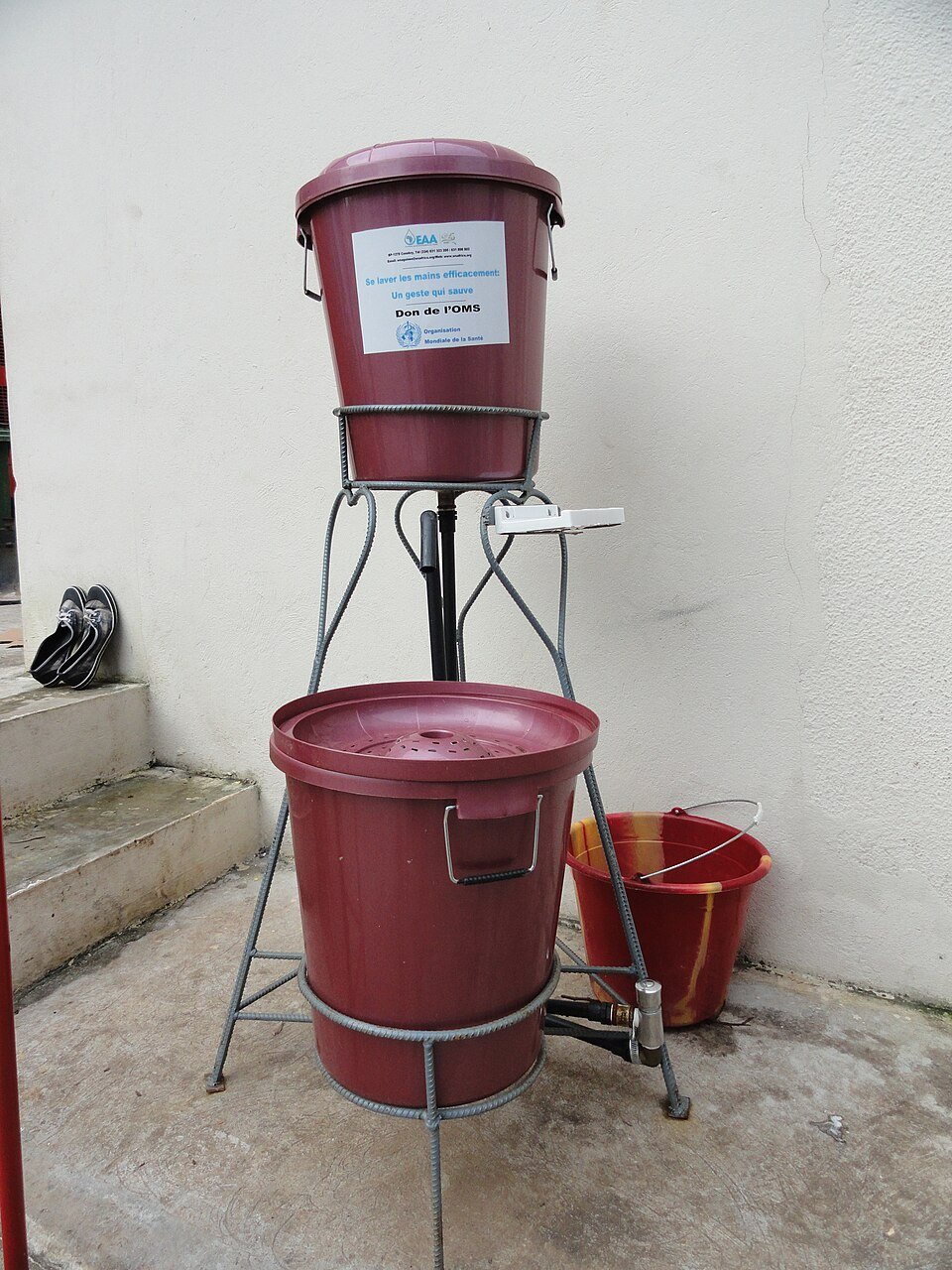A lidded bucket with a tap on a tall metal stand, with a second bucket below it to catch the water