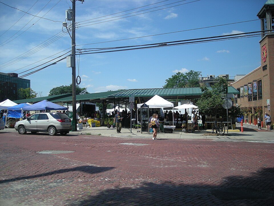 A market seen from across the street: white and blue pop-up canopies set up beside a permanent open shed