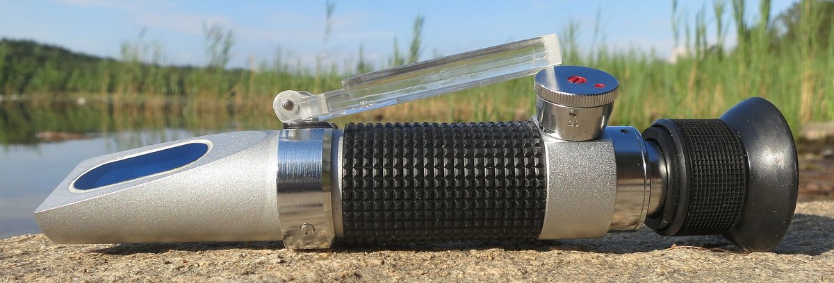 A handheld refractometer, a silver and black tube with an eyepiece and a hinged prism lid, lying on a stone by a lake