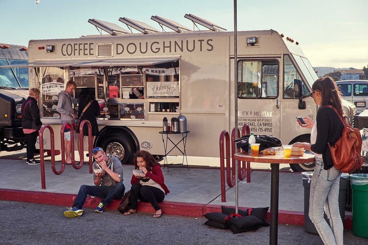 A beige food truck lettered COFFEE DOUGHNUTS with four solar panels on its roof and three airpots on a folding table beside the serving window, customers eating on the kerb