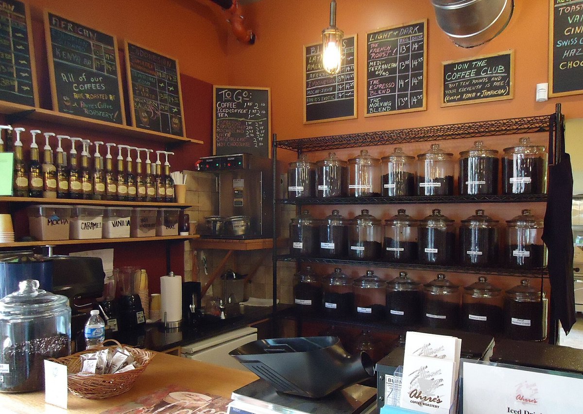 Inside a small roaster's shop: rows of glass jars of roasted beans on a wire rack, a scale and scoop on the counter, a batch brewer, flavour syrups on a shelf, and chalkboards listing coffees by origin and roast with a sign reading all of our coffees are roasted at Ahrre's Coffee Roastery