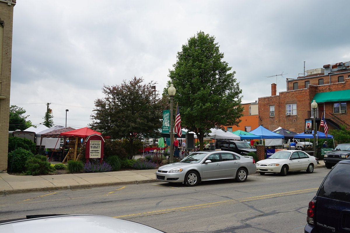 A small-town street with two parked cars in front of a row of white and blue canopies set up on a lot beside brick buildings
