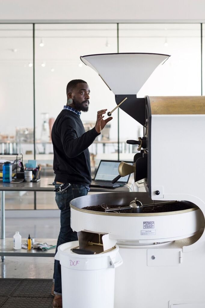 A roaster in a black sweater holds up the sample trier from a white production roaster, roasted beans turning in the cooling tray beside him and a laptop logging the roast behind