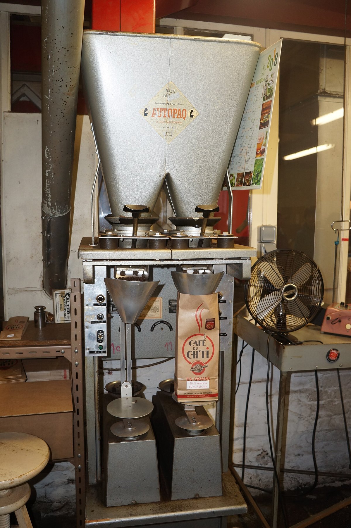 A white bag on the spout of a stainless weigh-filler hopper in a roastery, with labels and stock on shelves behind