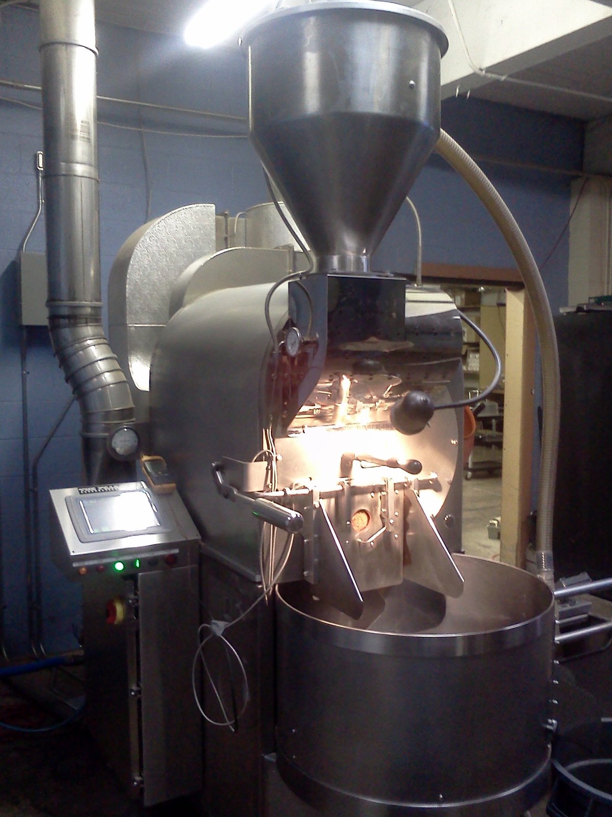 A tall black drum roaster with a hopper on top and a control screen, in a roastery with a white wall