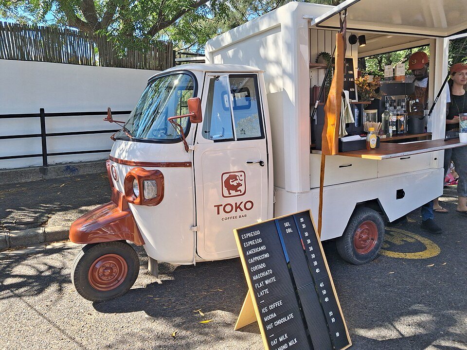 A white three-wheeled Piaggio van with its side raised to show an espresso machine, a menu board on the pavement beside it