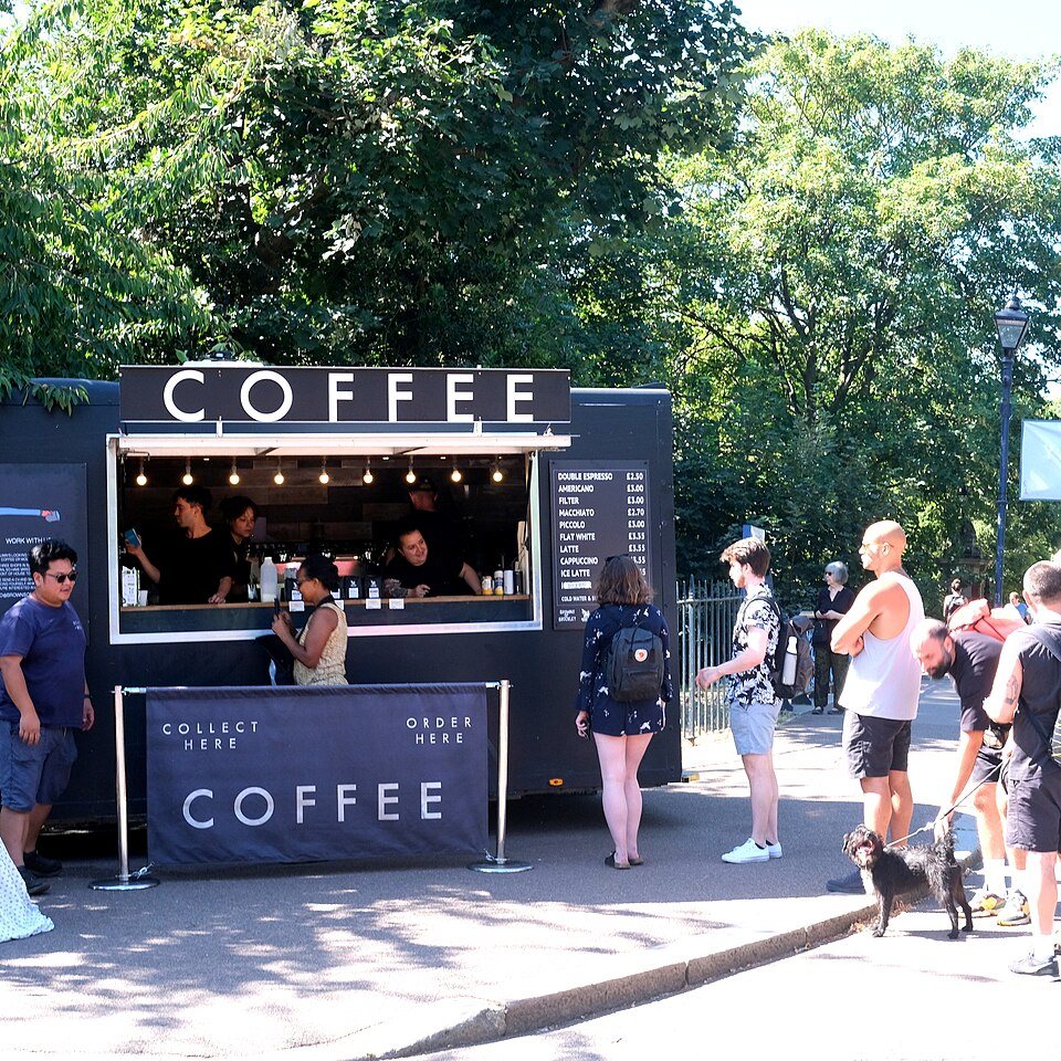 A dark box trailer with COFFEE in large letters over its open hatch, customers queuing in a park