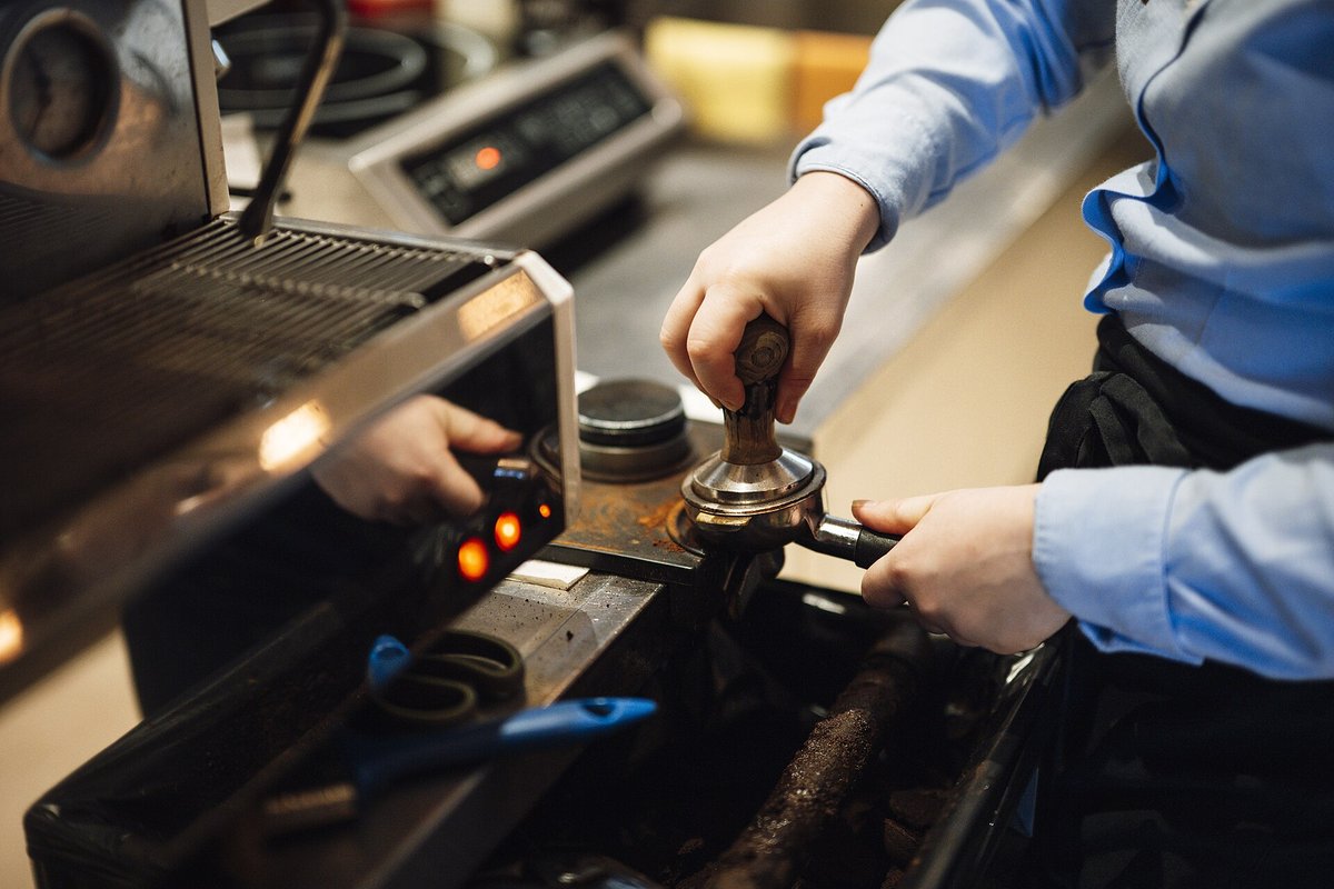 A barista in a blue shirt presses ground coffee into a portafilter with a tamper beside a commercial espresso machine