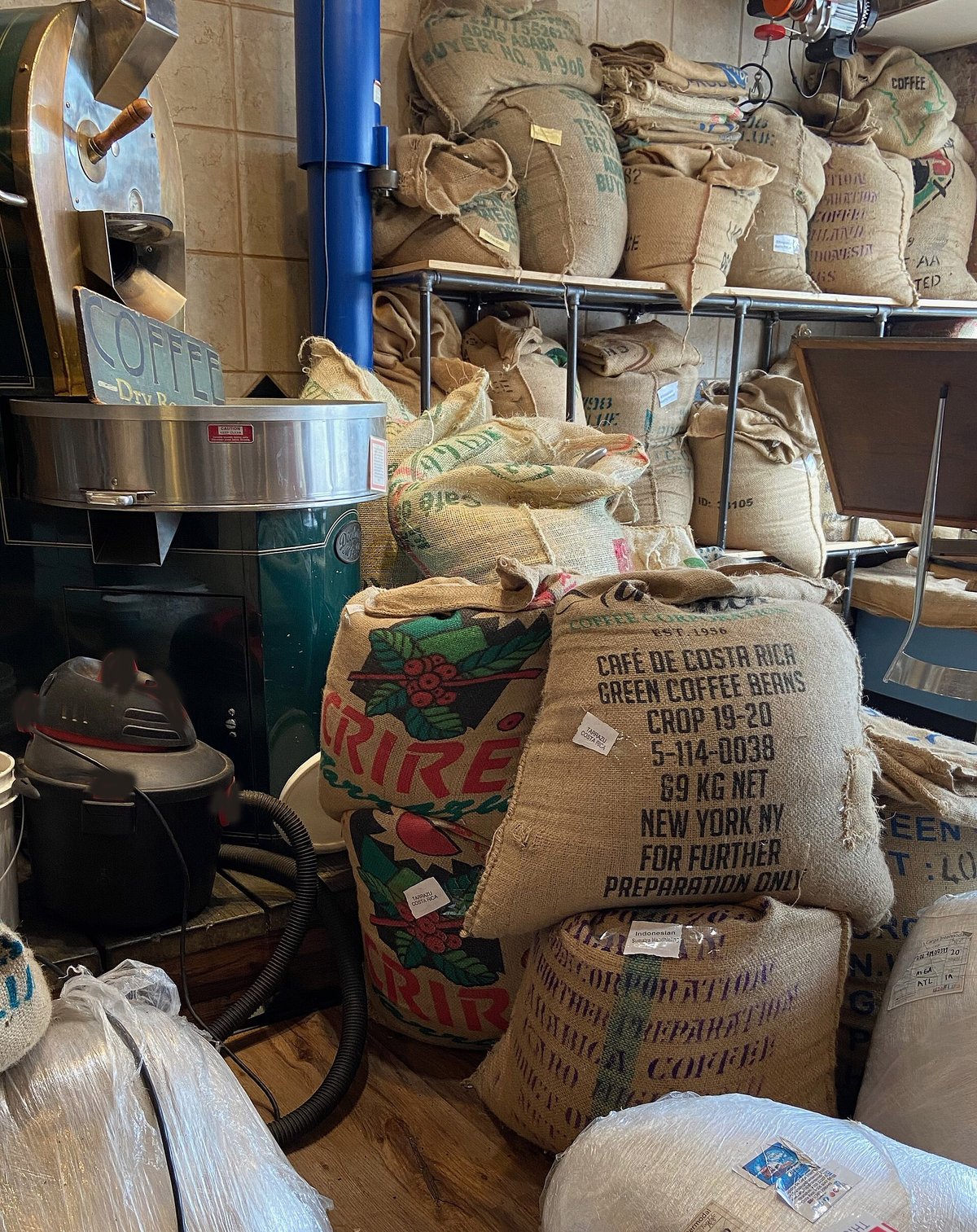 Inside a small coffee shop: a roaster, shelves and stacks of burlap coffee sacks printed with farm names