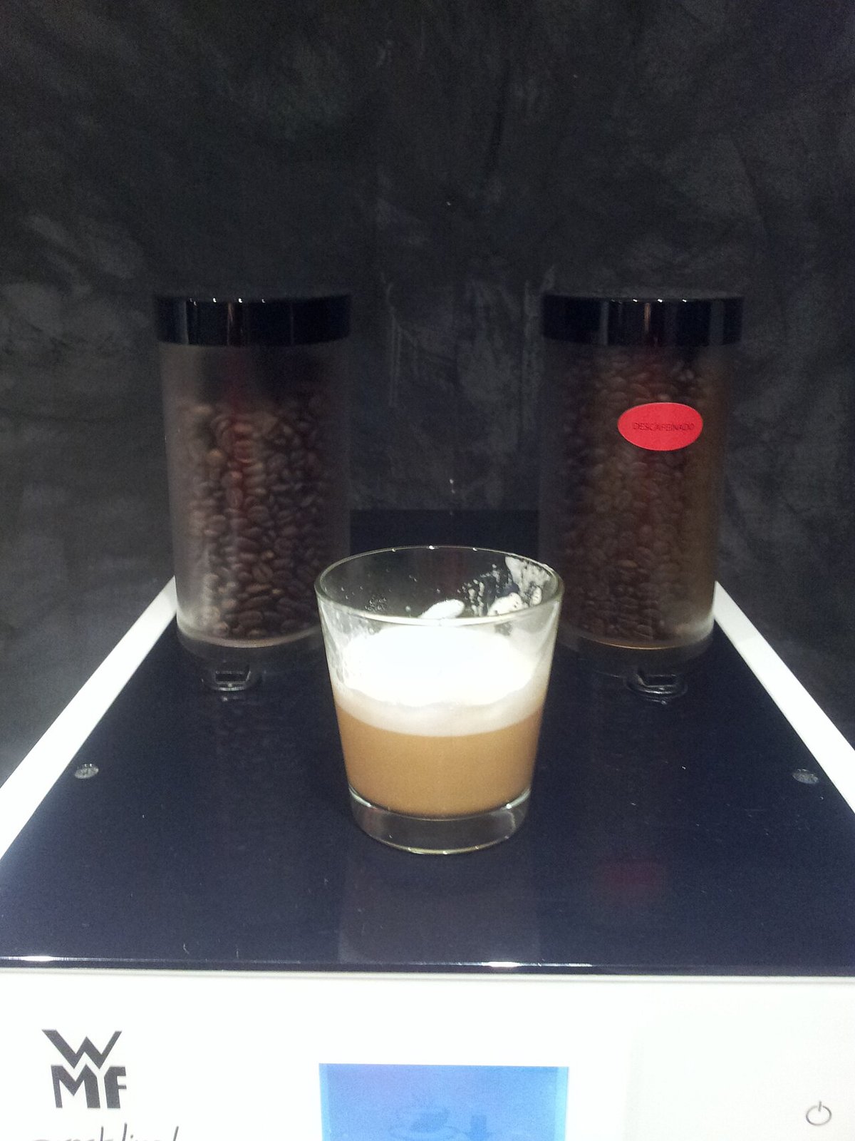 Two clear hoppers on a café espresso grinder, regular beans on the left and decaffeinated on the right, with a decaf cappuccino in front