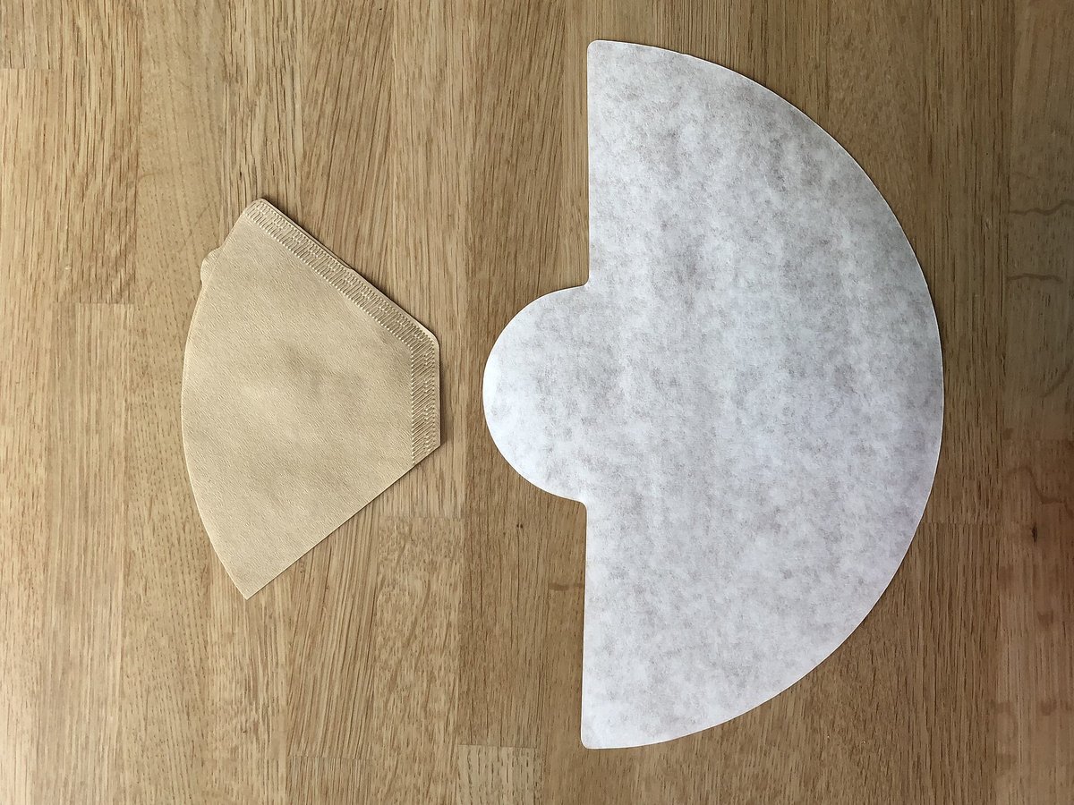 A cone of folded paper coffee filter on a wooden table beside a half-moon Chemex filter opened flat