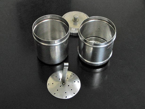 The parts of a South Indian stainless-steel coffee filter laid out on a dark counter: the upper perforated cup, the lower cup, the pressing disc with its handle, and the lid