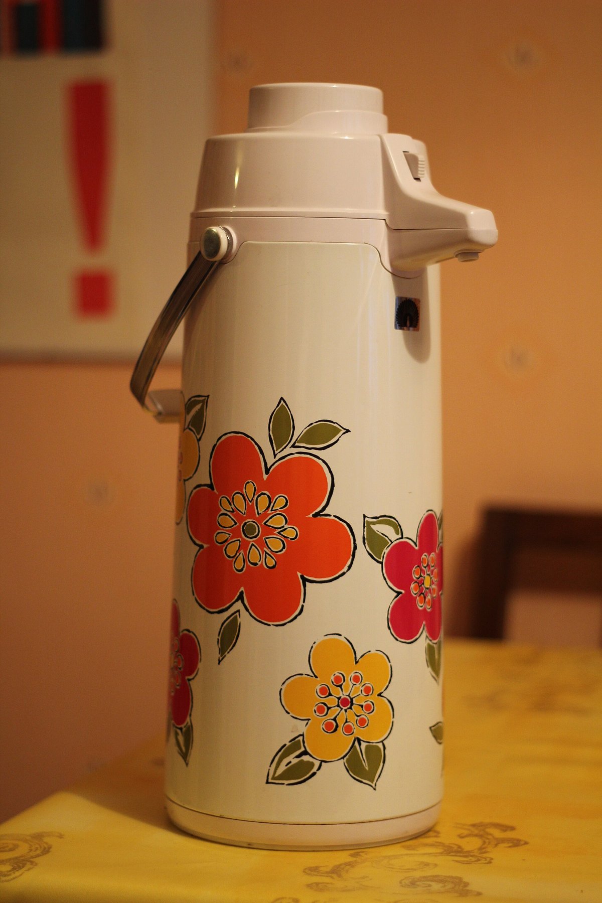 A pump-top vacuum airpot decorated with red and orange flowers, standing on a table