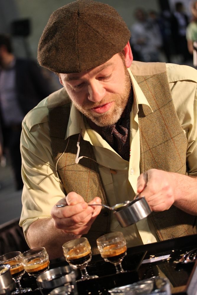 A competitor in a flat cap and waistcoat spoons an ingredient into a small steel pitcher above a row of glasses of espresso