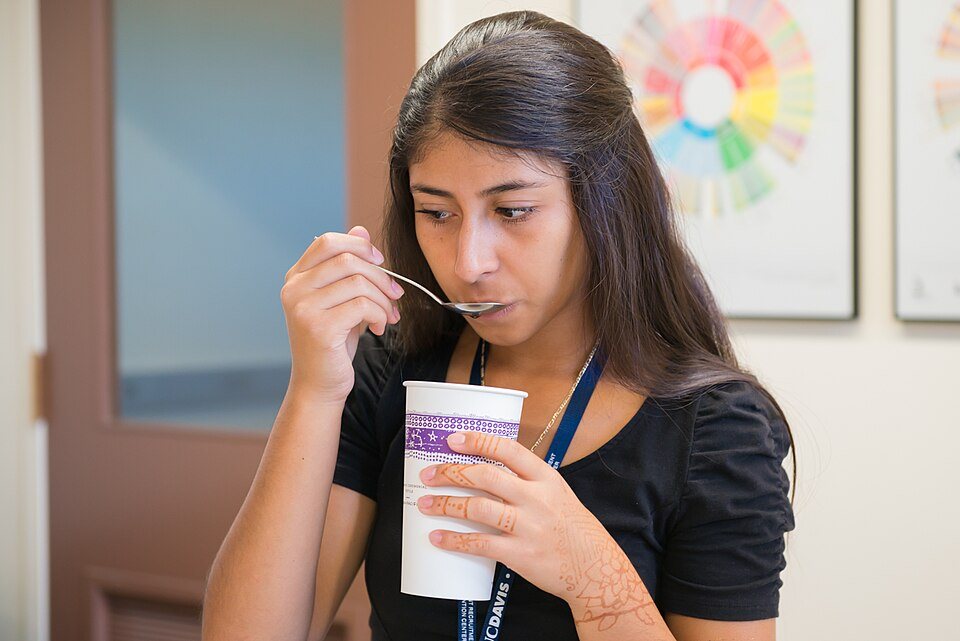 A student sips from a cupping spoon while holding a tall paper cup in her other hand, a coffee flavor wheel poster on the wall behind her