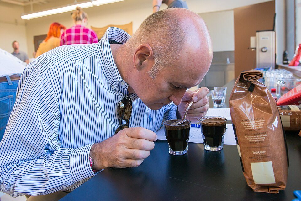 A man bends with his nose just above a glass of coffee and pushes a spoon through the cap of grounds