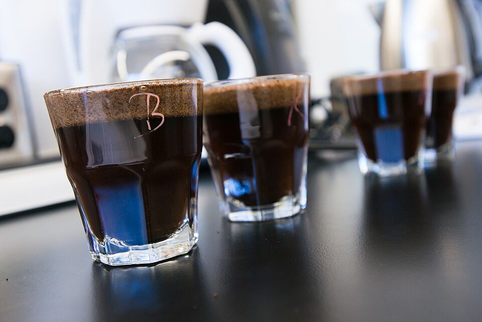 Four glasses of coffee on a black bench, each with a thick brown cap of wet grounds floating on top and a letter written on the glass