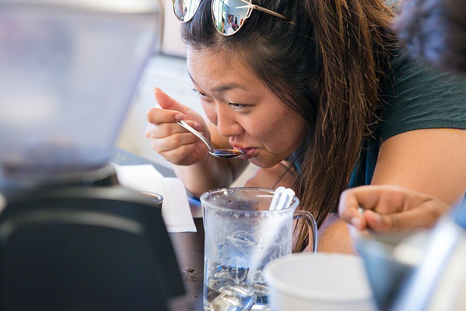A woman tastes from a spoon; in front of her stands a glass pitcher of water with spoons standing in it