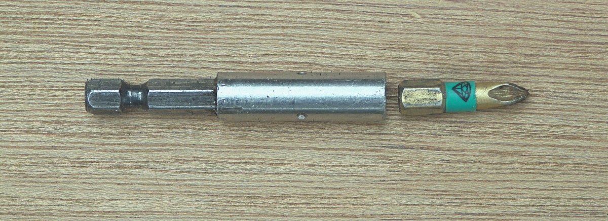 A steel screwdriver bit holder lying on a wooden surface: a six-sided shank at the left, a round silver barrel in the middle, and a Phillips bit seated in the barrel's socket at the right