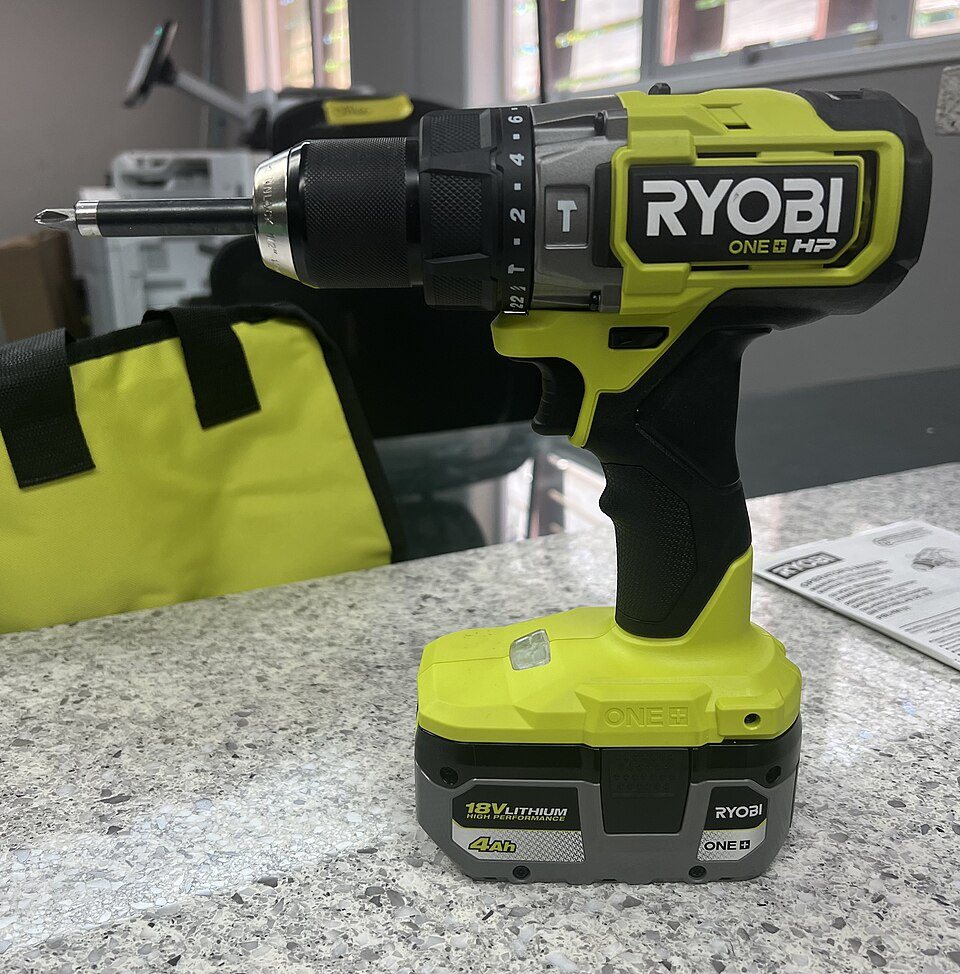 A yellow-green and black Ryobi ONE+ HP 18-volt cordless drill standing on its battery on a stone counter, a screwdriver bit in its chuck