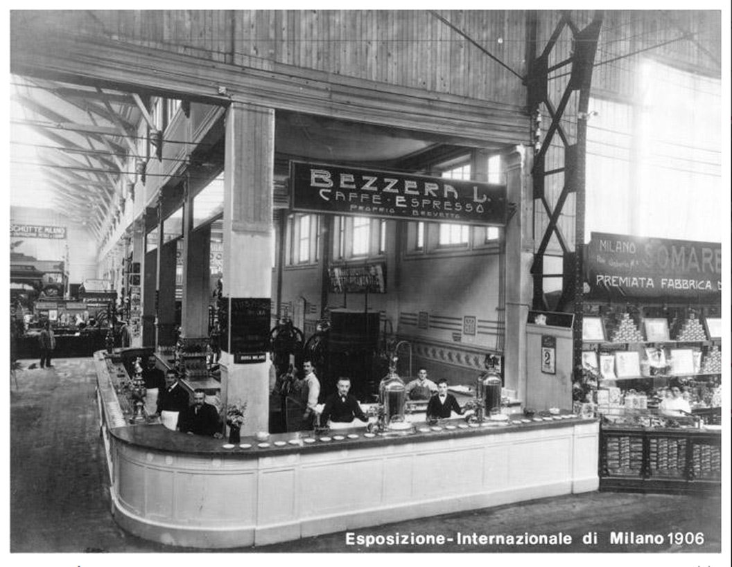 A long exhibition stand under a glass hall roof, with a curved counter and a sign reading Bezzera L. caffè espresso; a caption reads Esposizione Internazionale di Milano 1906