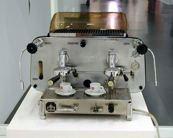 A two-group chrome espresso machine of 1961 on a plinth, with a smoked-glass cup tray on top, two cups under the groups and the Faema name on the front