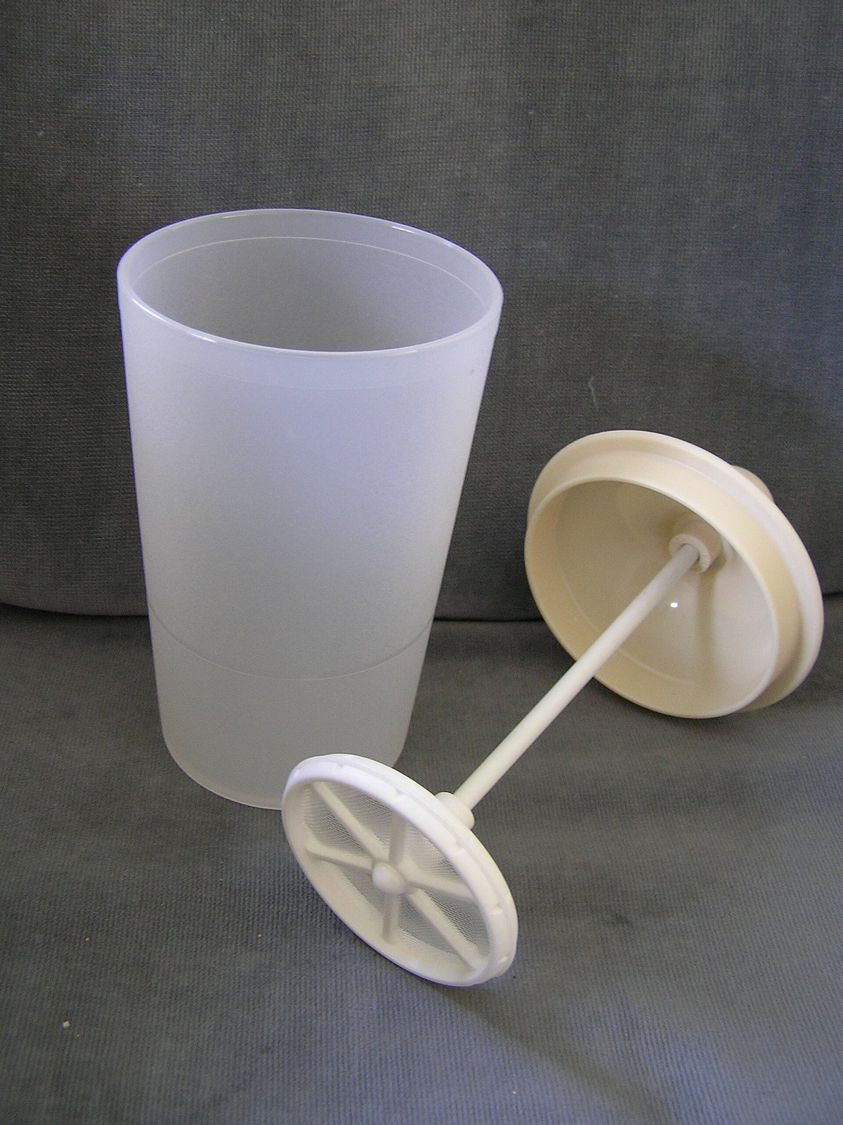 A tall frosted plastic jug beside its lid, with a plunger rod ending in a round mesh disc lying in front of it