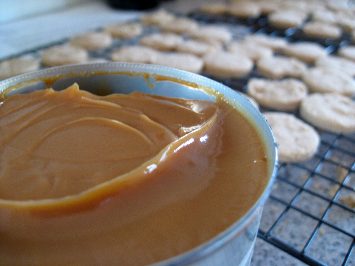 A metal pot of thick, glossy brown dulce de leche with cookies cooling on a rack behind