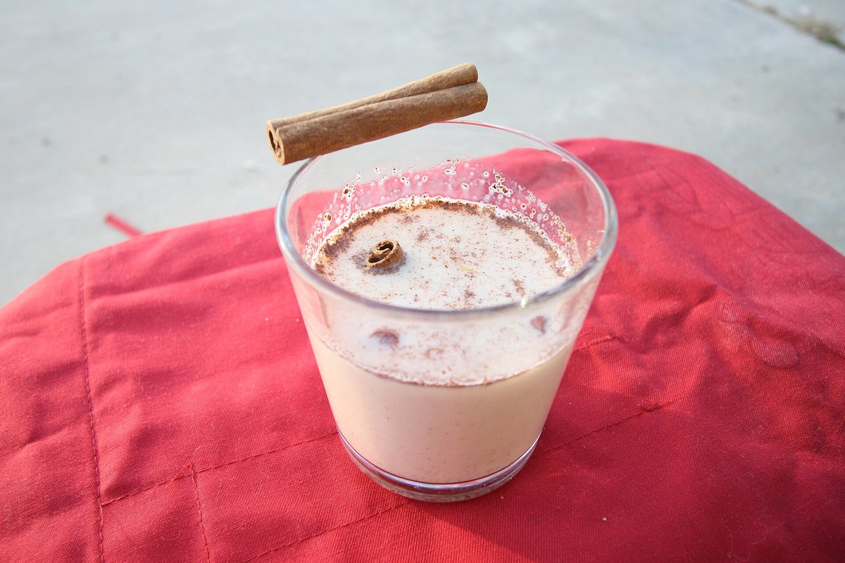 A clear glass of pale eggnog dusted with cinnamon, a cinnamon stick laid across the top, on a red cloth