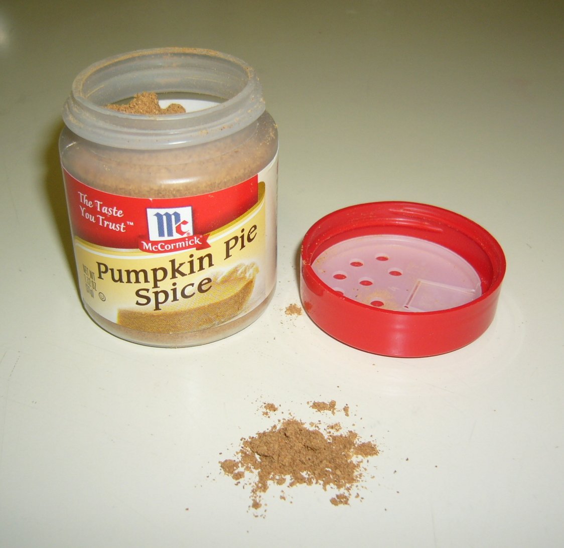An opened jar of ground pumpkin pie spice beside its red shaker lid, with a little spice spilled on the table