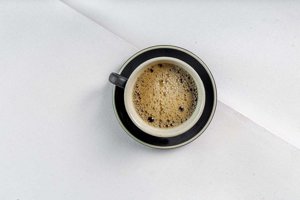 An espresso in a white cup on a black saucer, seen from above, with a mottled tan crema covering the surface