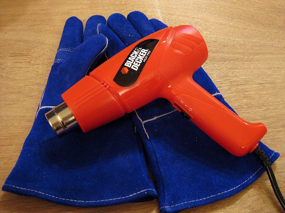 An orange heat gun lying on a pair of blue leather welding gloves