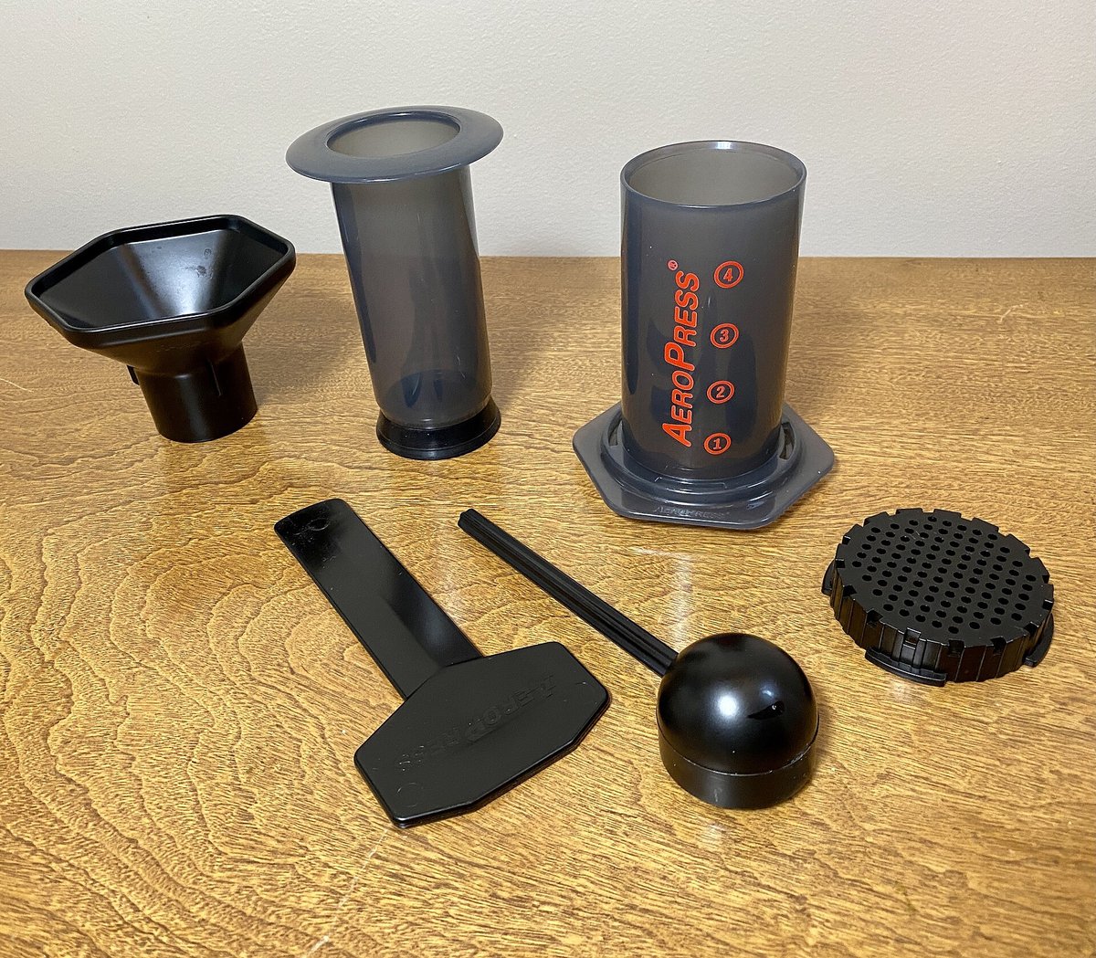 A red-lettered AeroPress with its funnel, scoop, stirrer, filter cap and plunger laid out on a wooden counter